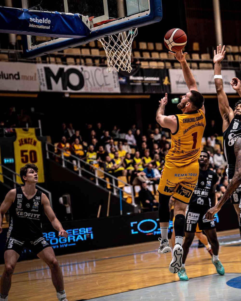 Basketball player in yellow jersey #1 drives to the hoop for a layup during an indoor professional league game.