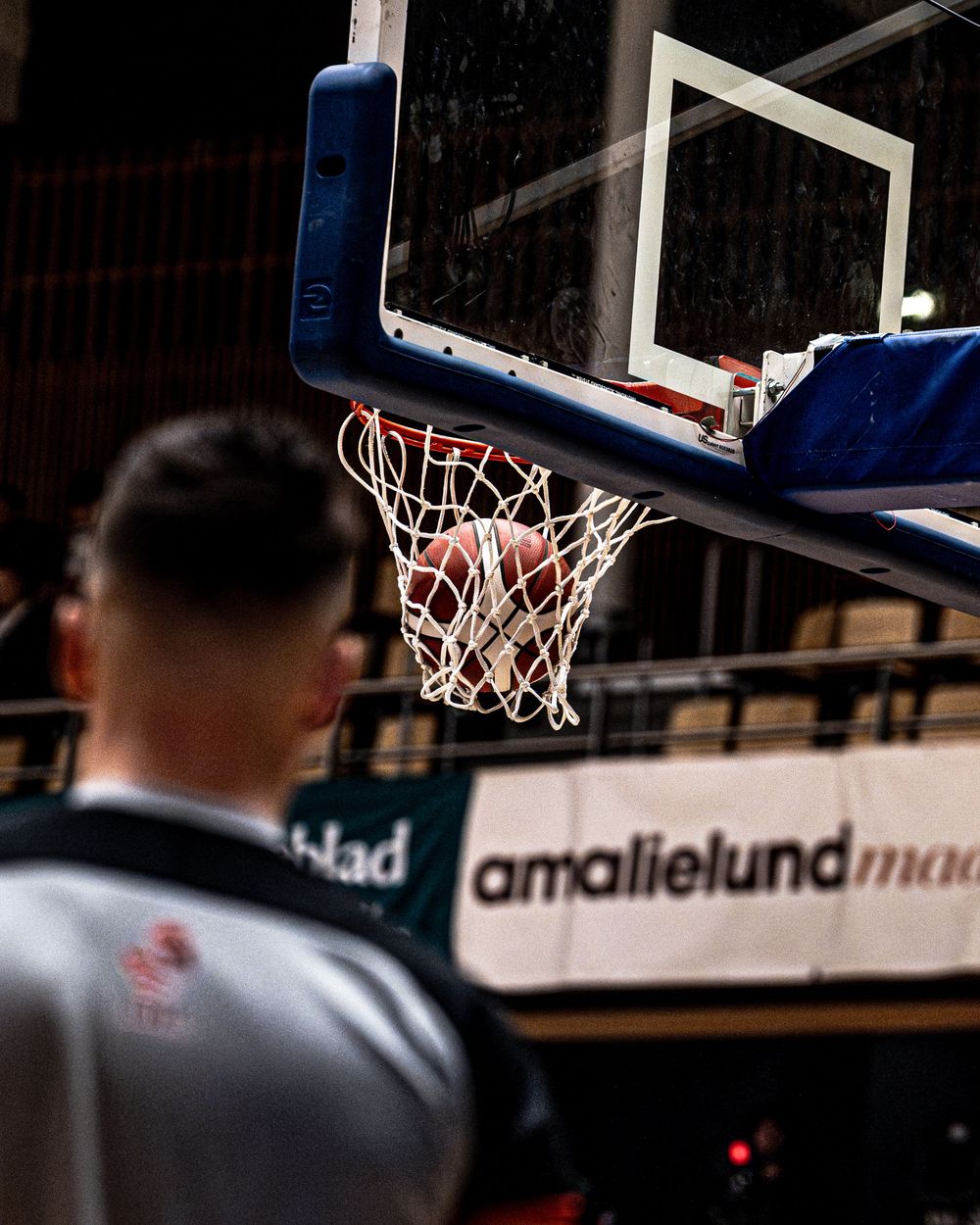 Basketball passing through the hoop net during a game, with a player in foreground and amalielund advertisement visible.