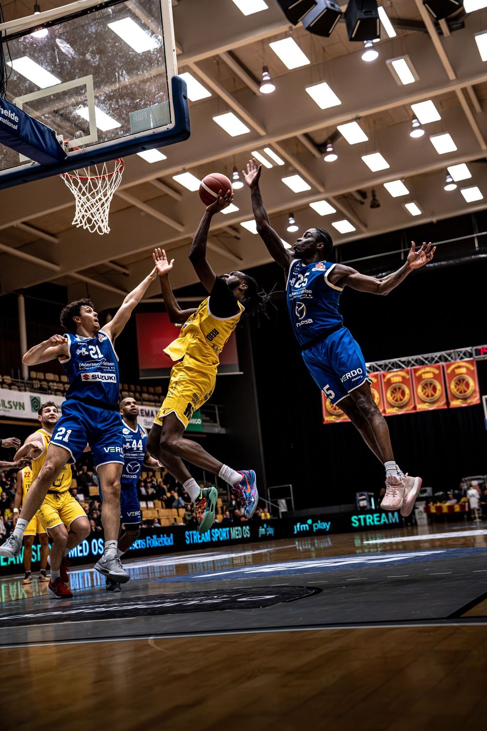 Basketball player in yellow drives to the hoop against blue-uniformed defenders in an indoor arena game.