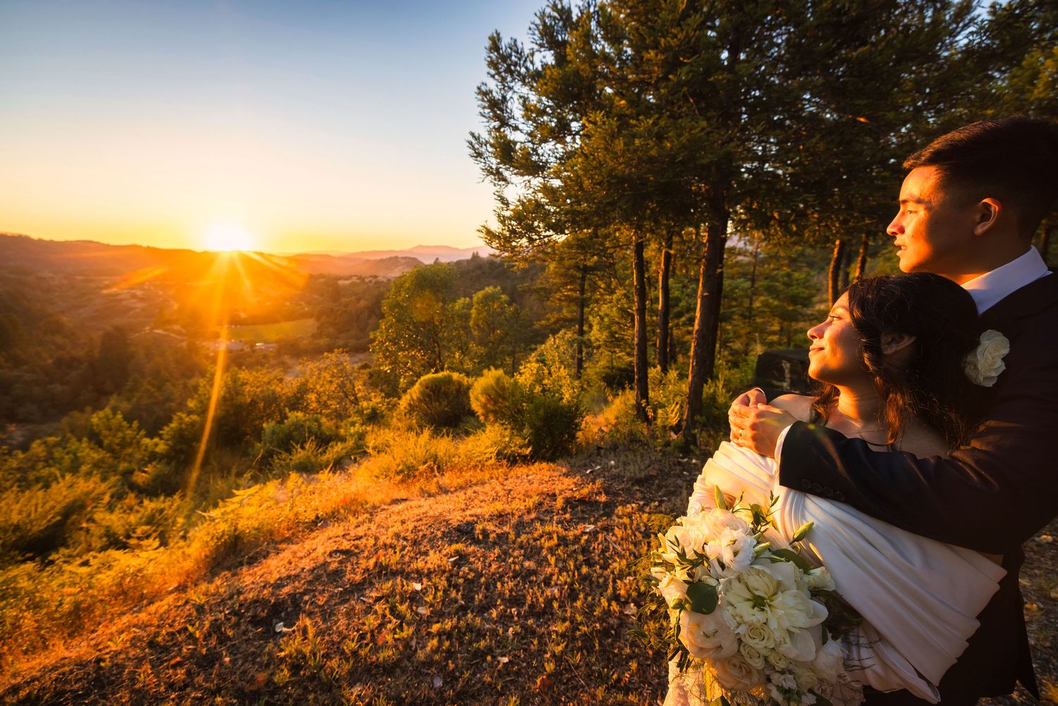 Bride and groom embrace at golden sunset overlooking scenic valley, surrounded by pine trees on rocky hilltop.