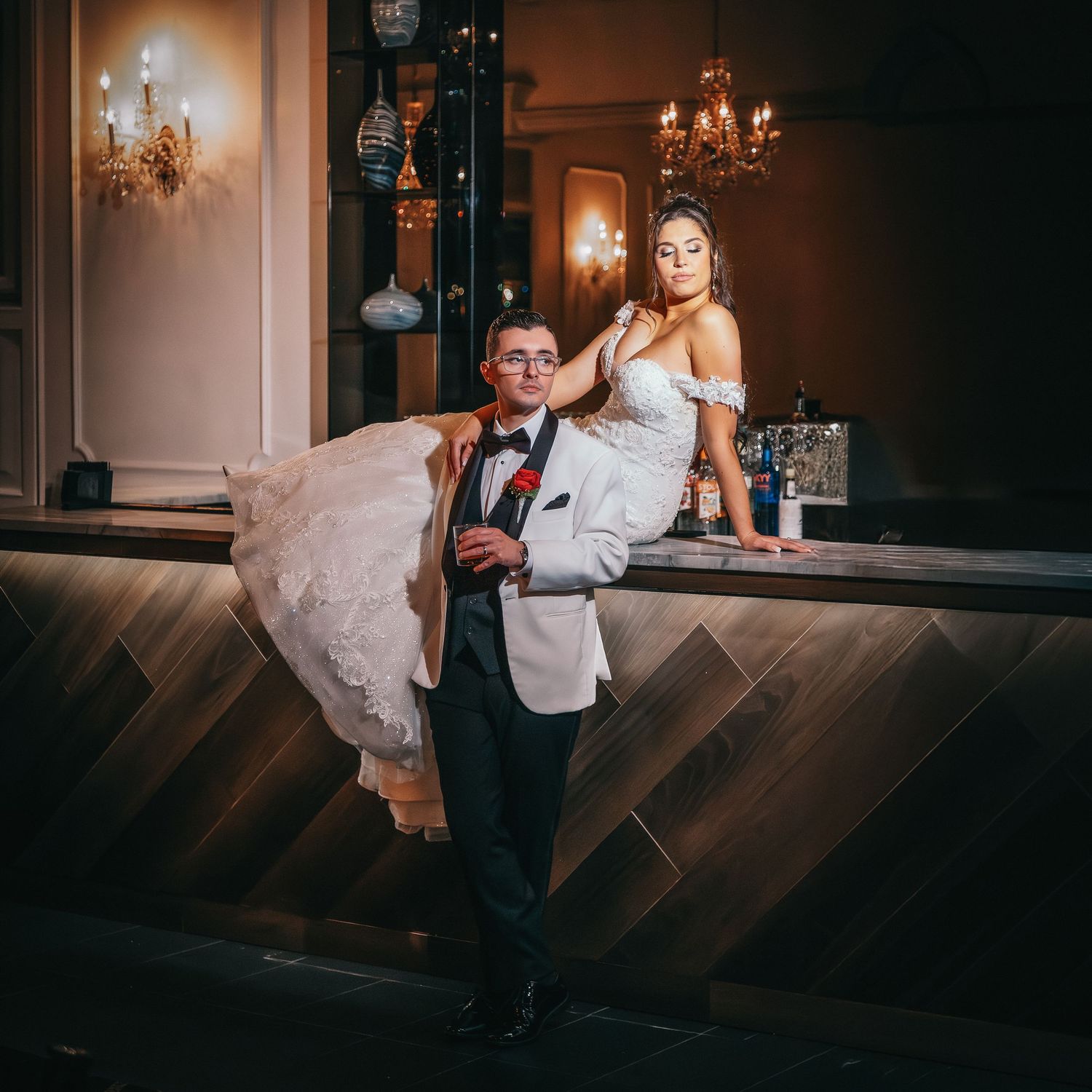 A bride in a white gown leans on a bar while a groom in a white tuxedo jacket holds a rose in an elegant venue.
