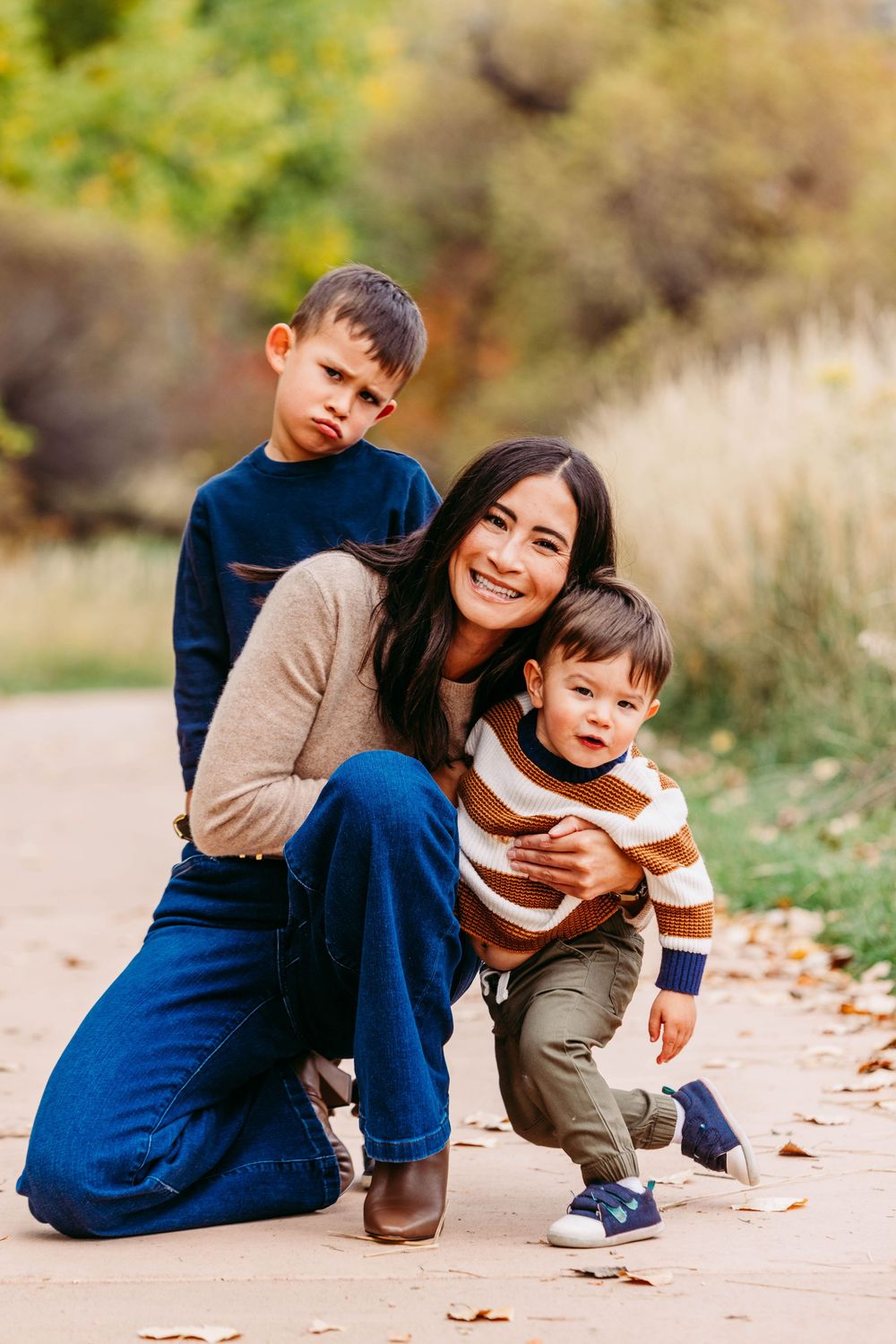 A smiling mother crouches with her two young sons on a leaf-covered path during autumn.