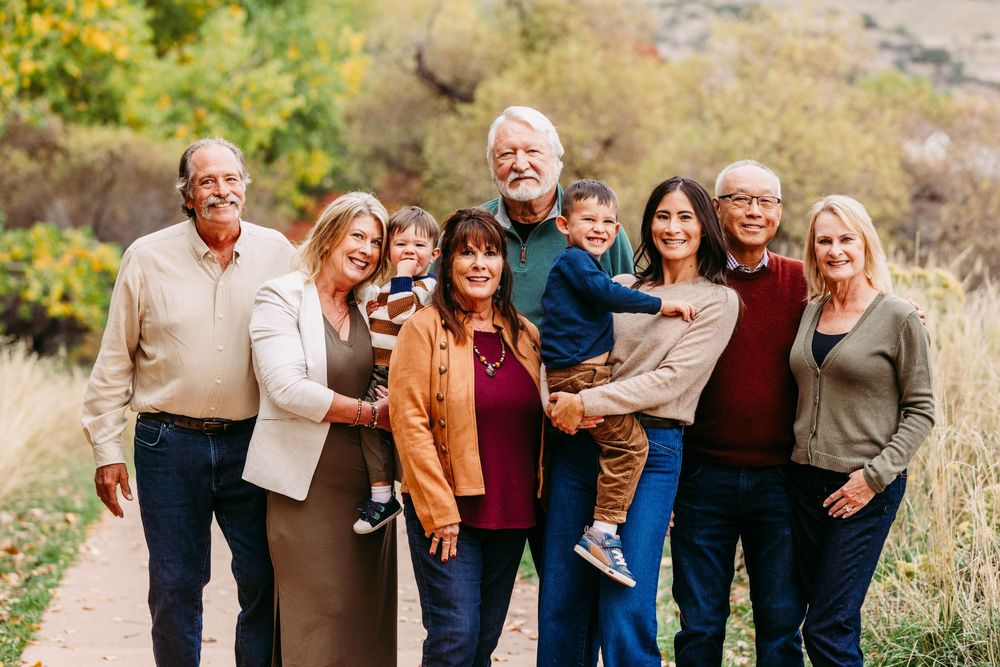 Multi-generational family smiling together outdoors on a scenic autumn trail with colorful foliage in the background.