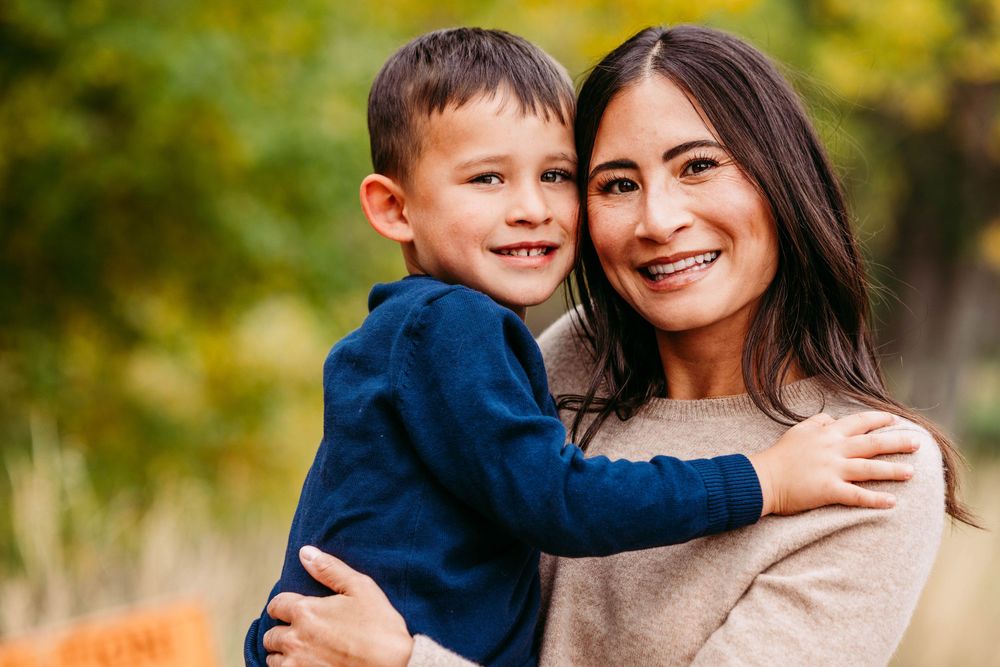 A smiling woman and young boy share a warm hug outdoors, surrounded by autumn foliage in a family portrait.