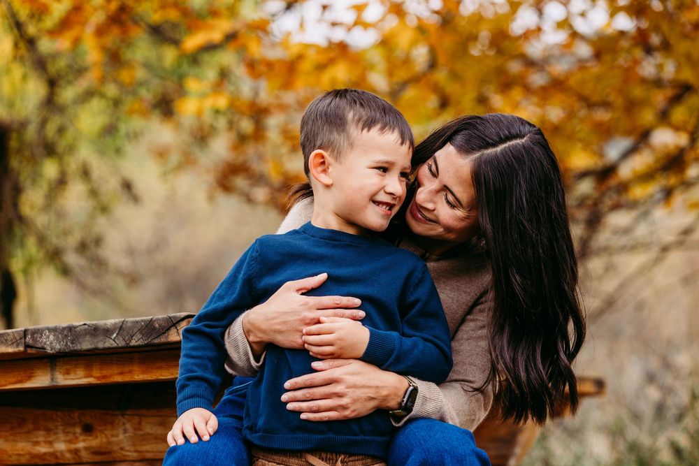 A mother hugs her smiling toddler son on a wooden bench surrounded by colorful autumn foliage.