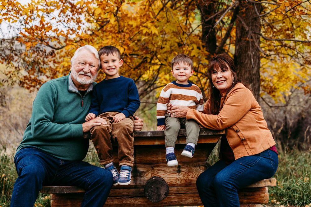 Family of four posing outdoors in autumn, two children sitting on wooden bench with grandparents beside colorful fall foliage.
