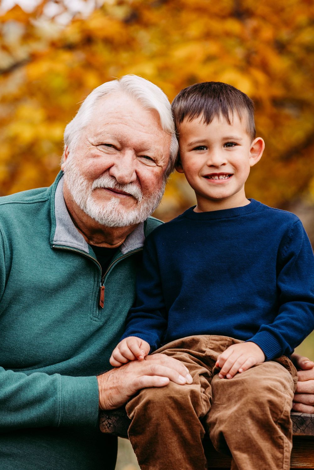 Elderly grandfather with white beard holding smiling grandson outdoors with fall foliage background.