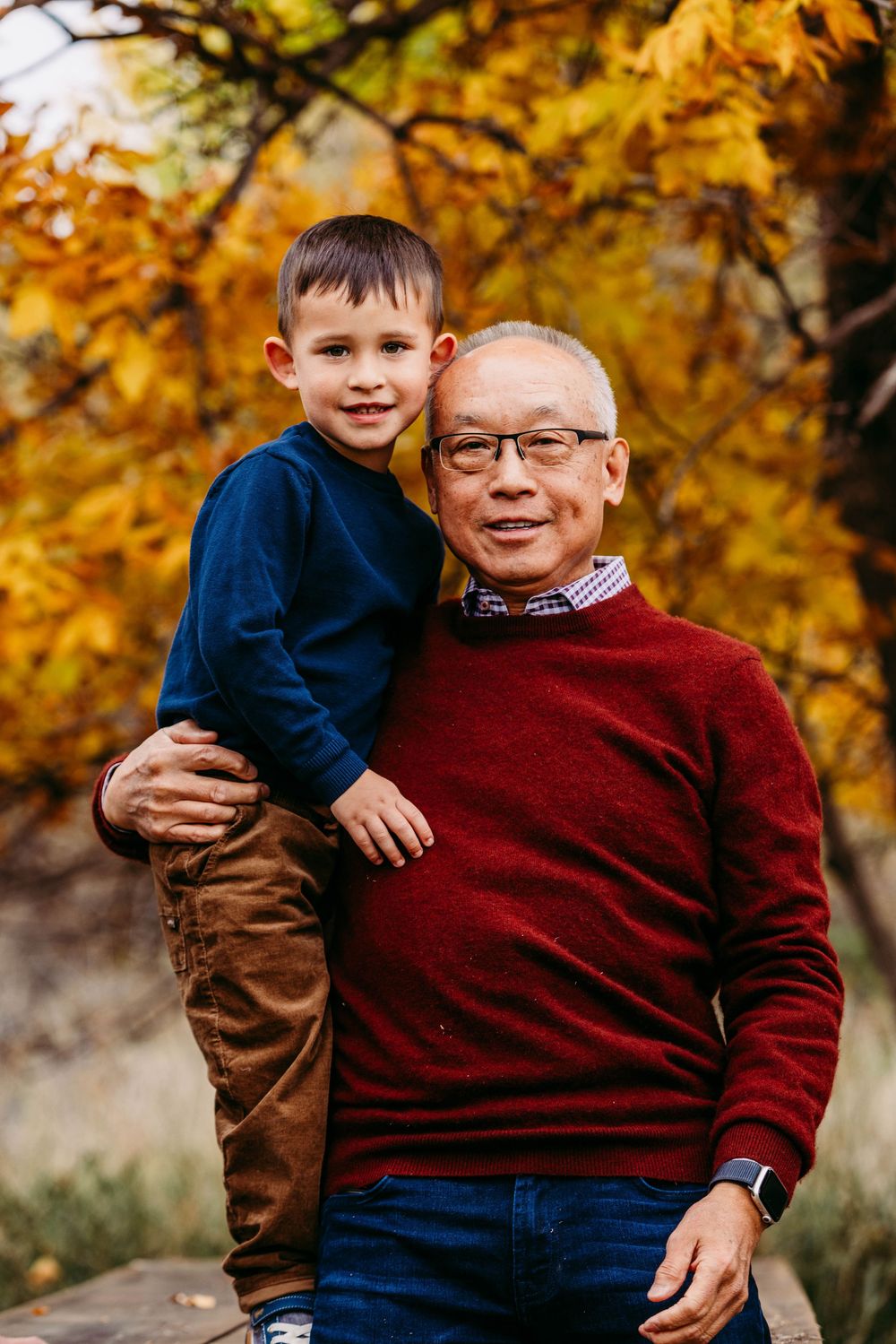 Grandfather in red sweater holds young grandson outdoors amid vibrant autumn foliage.