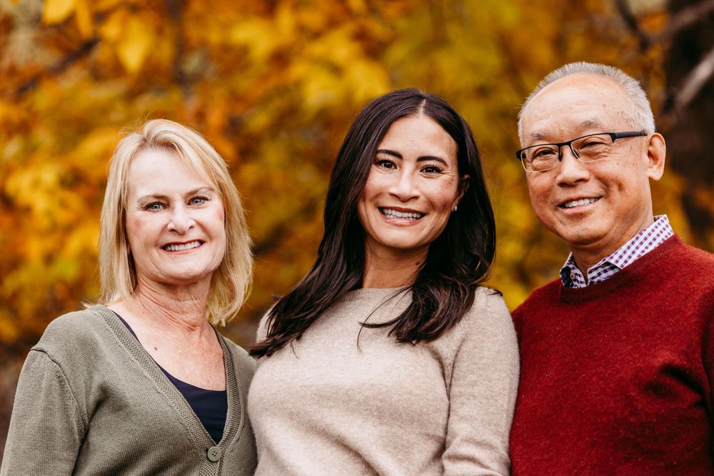 Three smiling adults pose together outdoors among vibrant autumn foliage in warm fall colors.