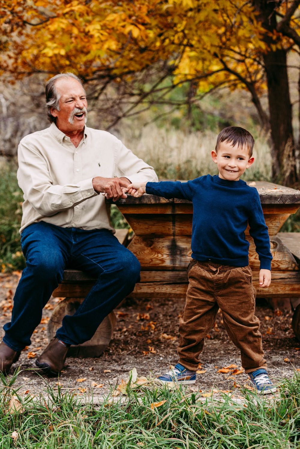 Grandfather sitting on bench laughing while holding hands with young grandson in autumn park.