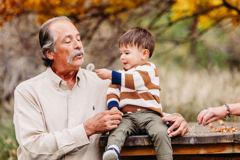 Grandfather sitting on bench with young toddler grandson on his lap during autumn family photo session outdoors.