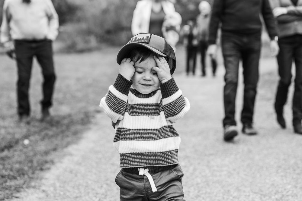 A young boy in a striped sweater and cap covers his eyes while walking on a path outdoors.
