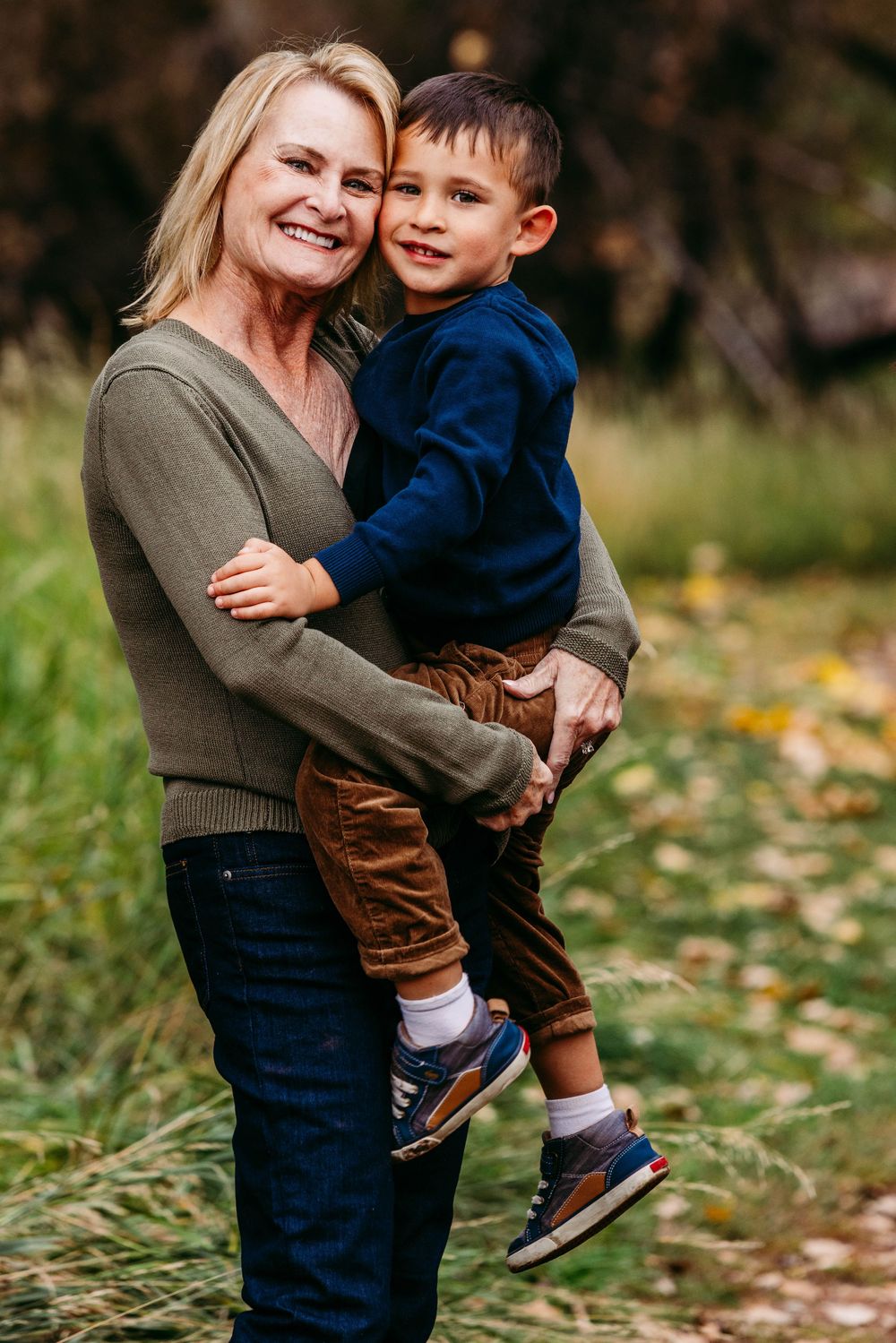 Smiling blonde woman holding a young boy in a blue sweater outdoors in an autumn park setting.