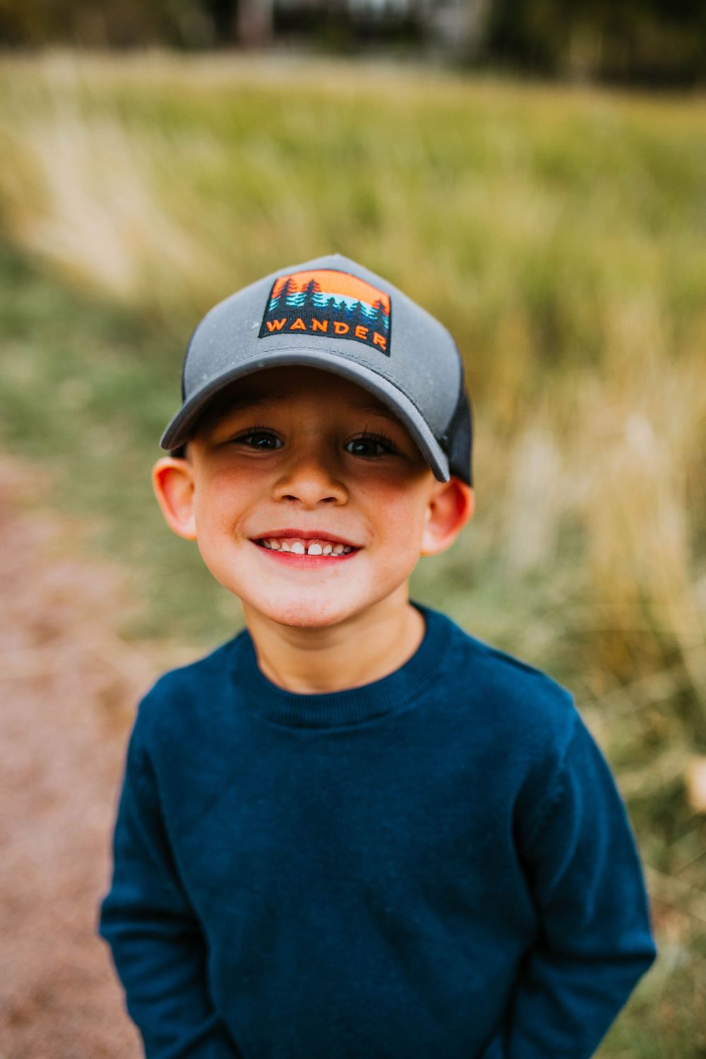 Smiling young boy wearing a gray Wander trucker hat and navy blue sweater outdoors in a field.