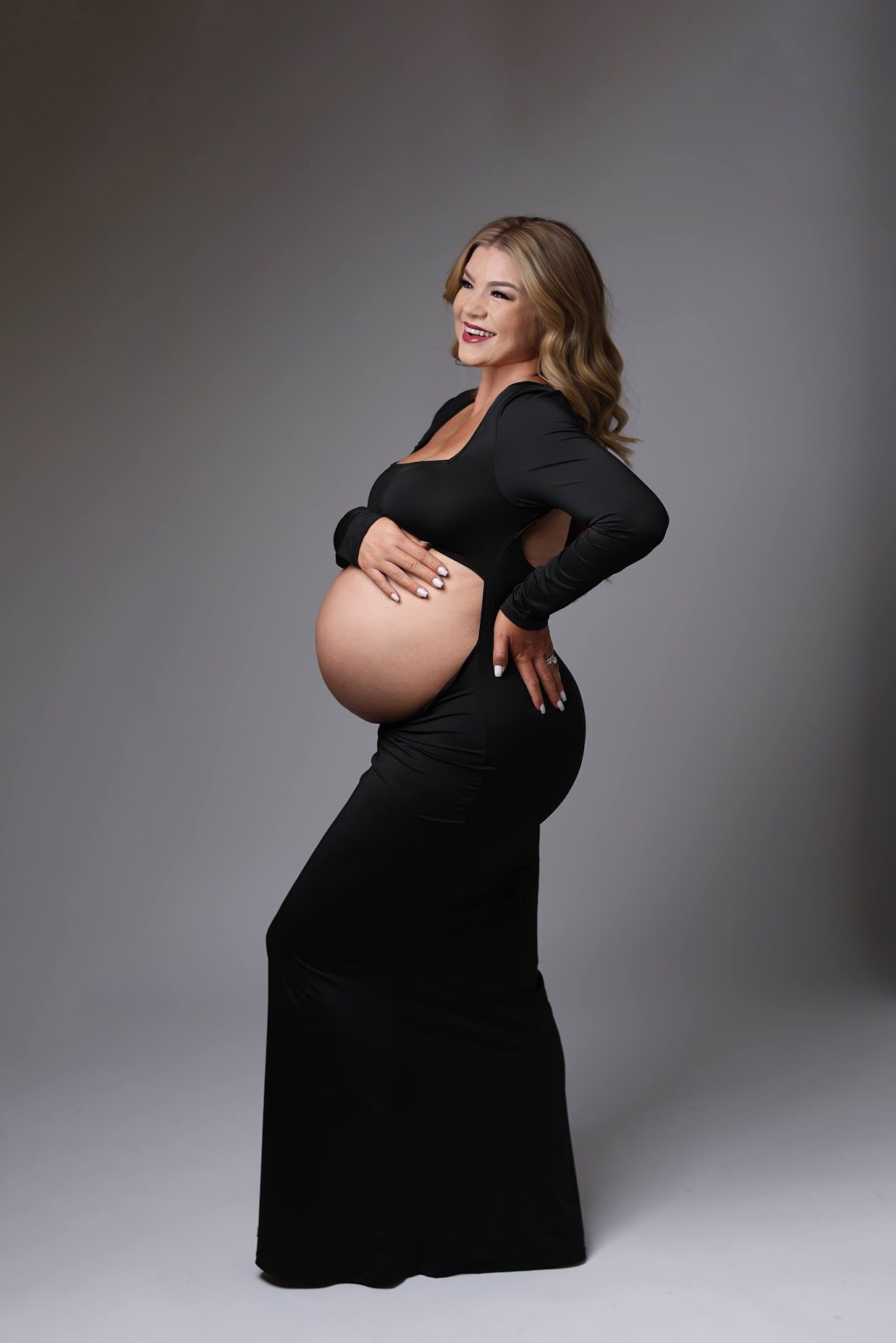 A pregnant woman in a black form-fitting gown poses sideways, cradling her bare baby bump in a studio.