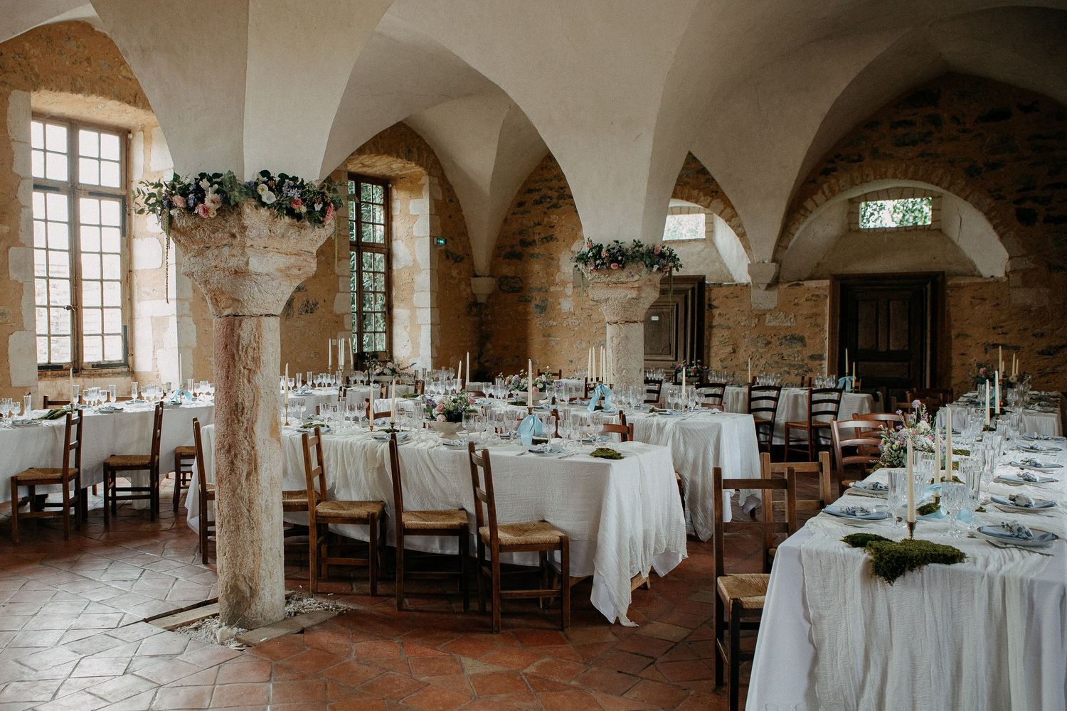 Elegant wedding reception in a rustic stone vaulted hall with white tablecloths, wooden chairs, and floral arrangements.