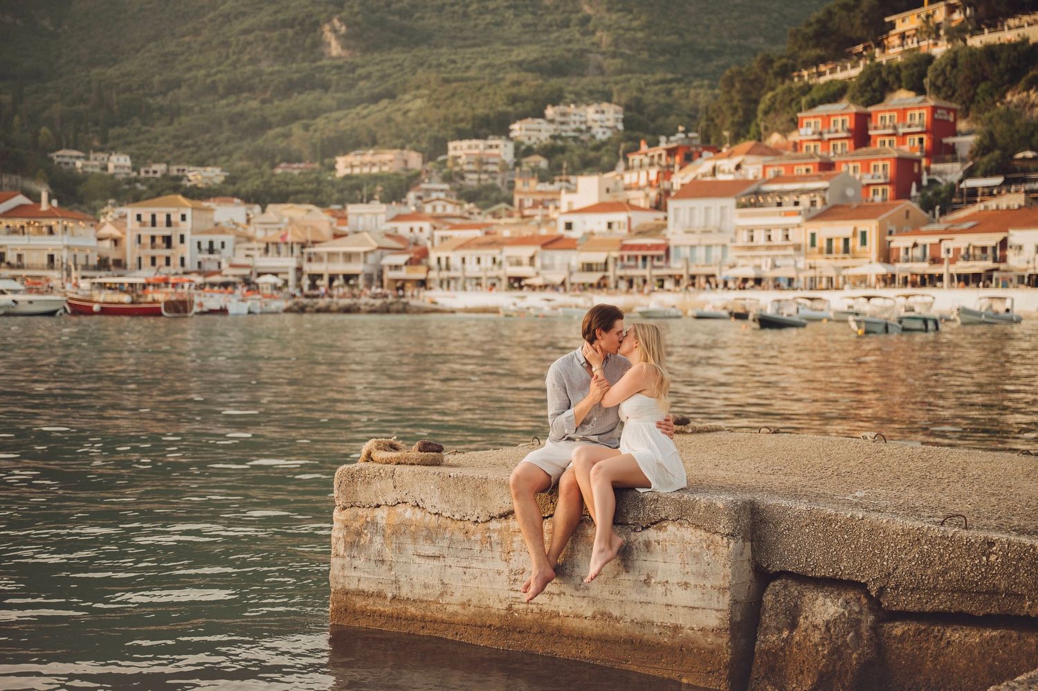 Couple sitting on a stone pier overlooking a picturesque Mediterranean coastal town with colorful buildings and green hills.