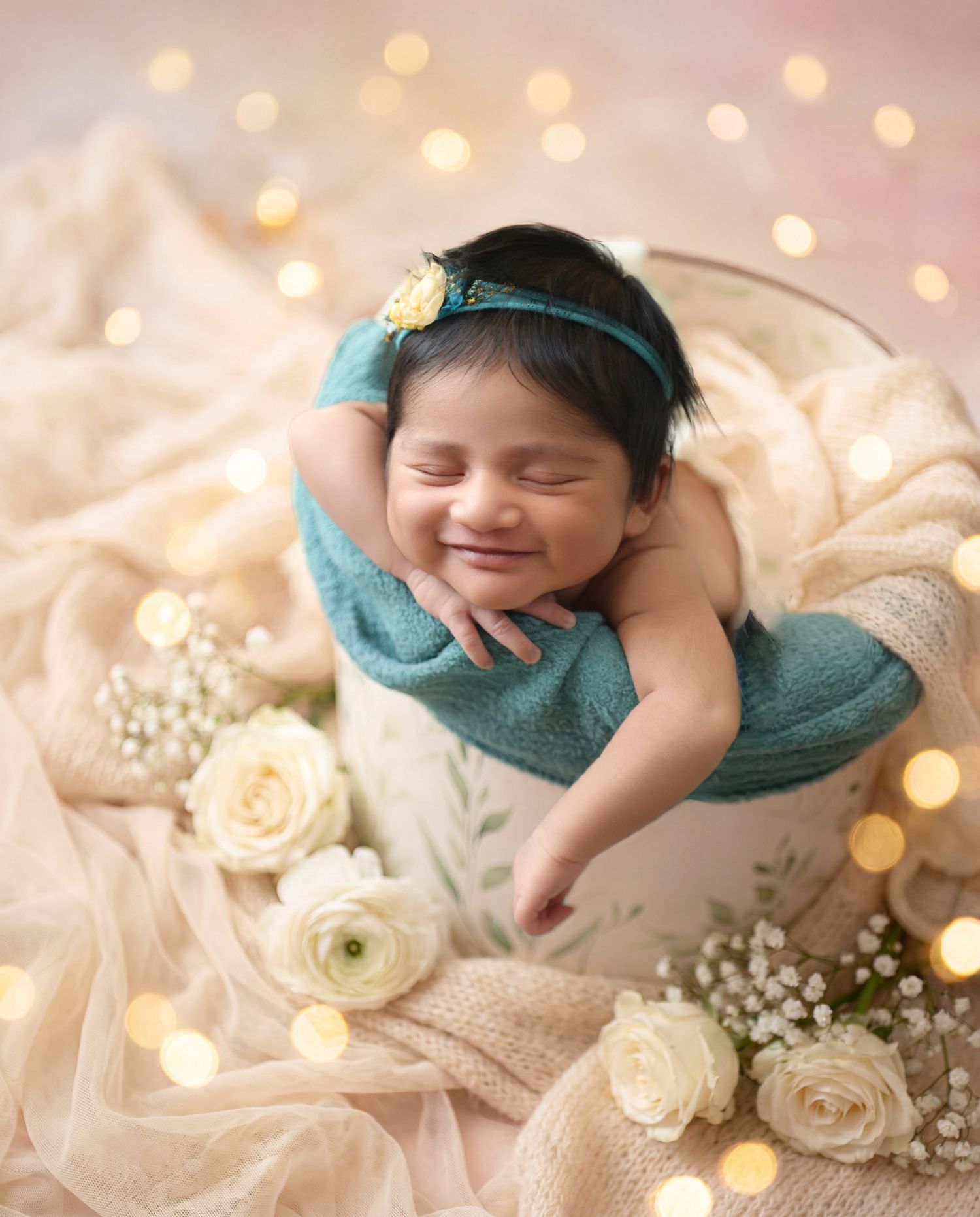 Smiling newborn baby wrapped in teal cloth sitting in a white bucket surrounded by white roses and bokeh lights.