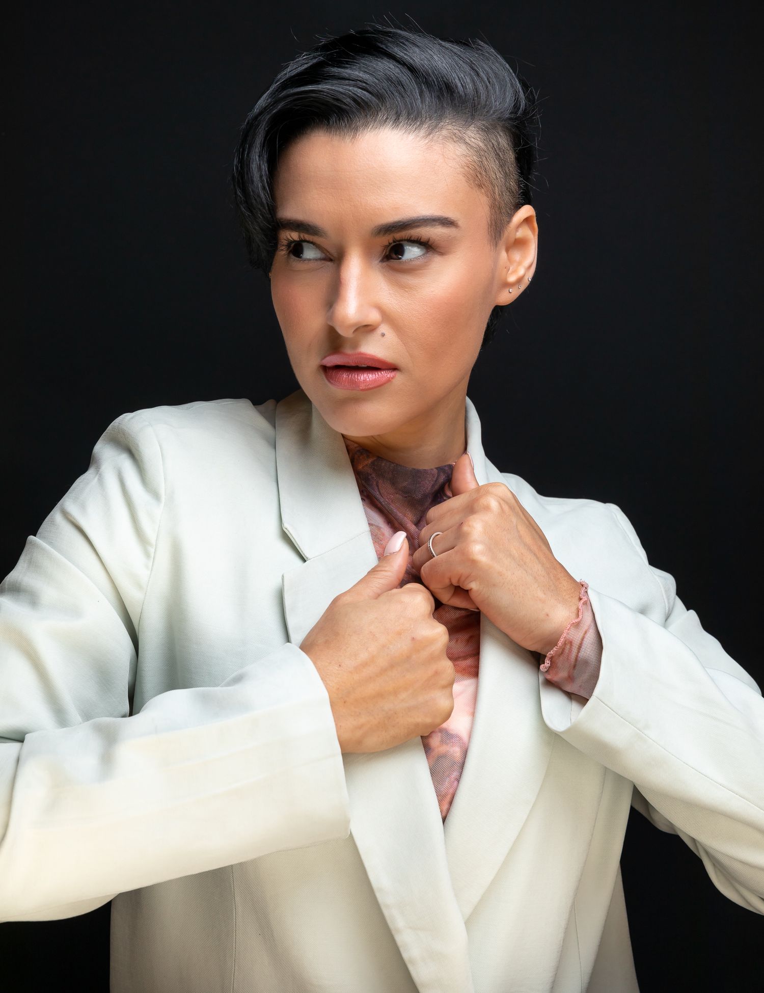 Woman in white blazer with short undercut hairstyle poses confidently against dark background.