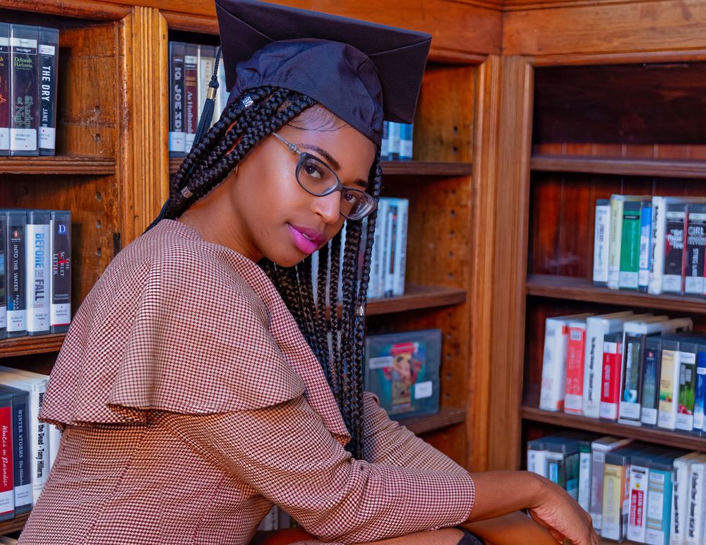Graduate in cap and gown poses by library bookshelves wearing glasses and pink blouse.