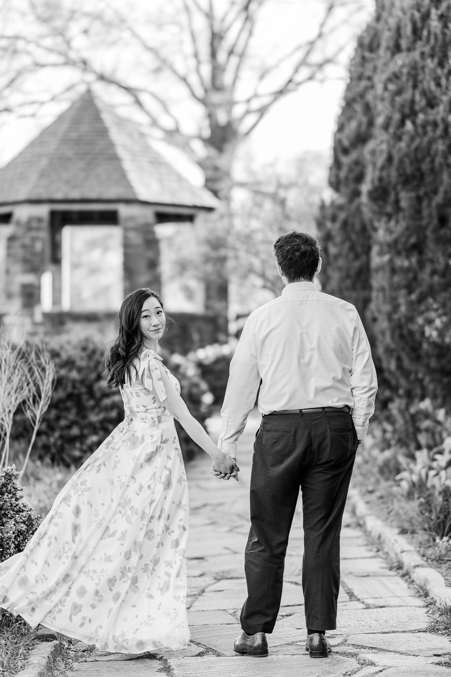 Black and white photo of a couple holding hands walking along a garden path toward a gazebo.