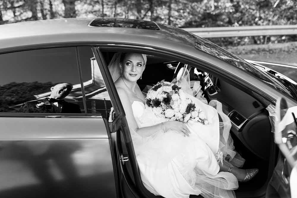 Bride in white wedding dress holding bouquet sits in car door in black and white photo.
