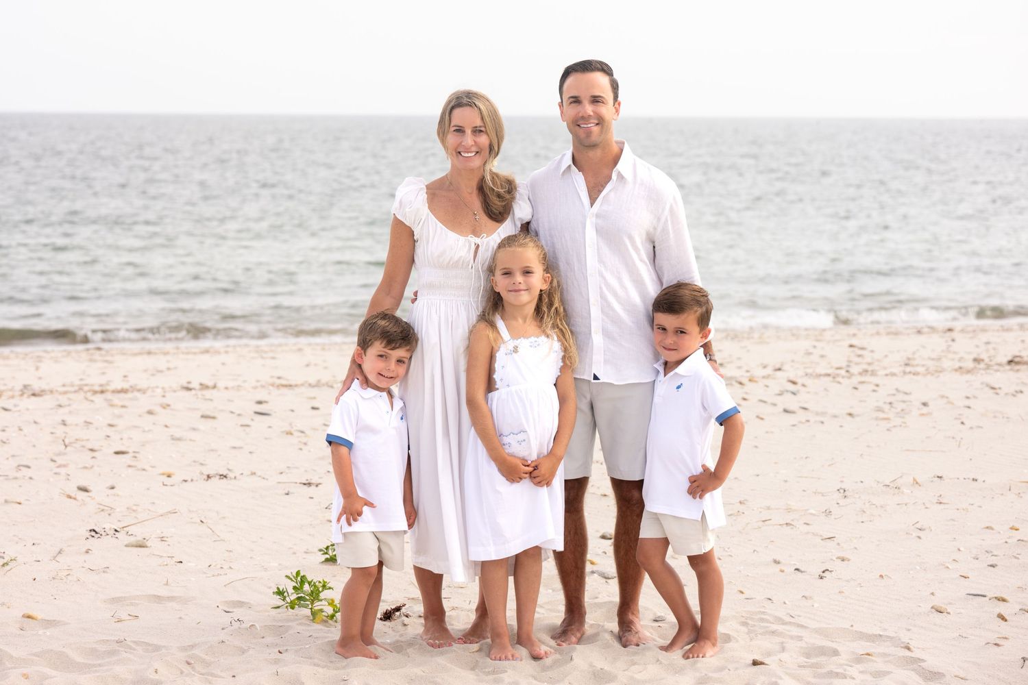Family of five posing on a sandy beach, parents and three young children dressed in white and khaki outfits.