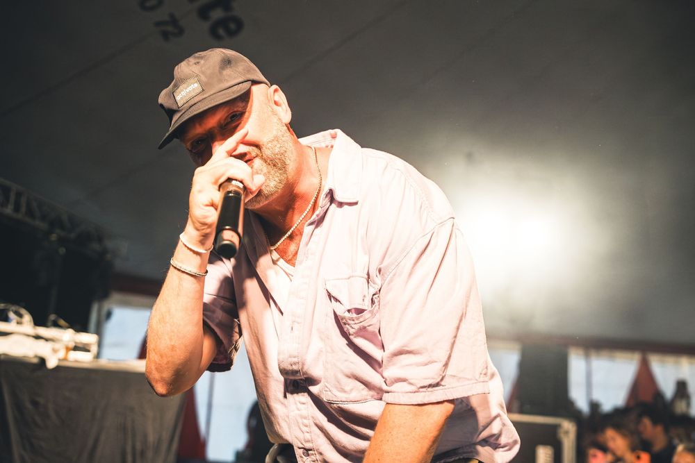 Man singing passionately into microphone on stage at outdoor music festival, wearing cap and pink shirt.