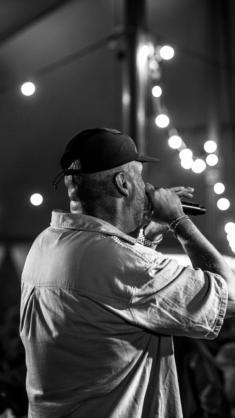 Black and white photo of a male rapper performing on stage, holding a microphone, wearing a cap and chain necklace.