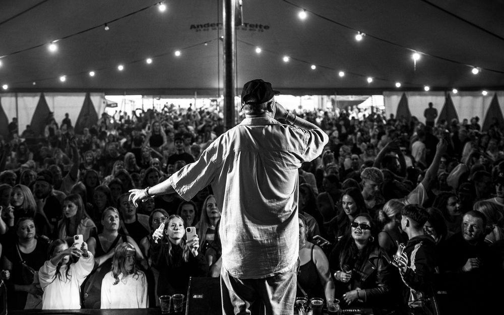 Black and white photo of a performer on stage facing a large crowd under a tent with string lights.