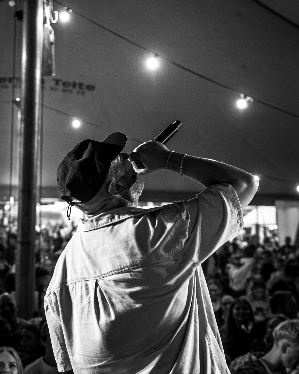 Black and white photo of a performer holding a microphone to a crowd under a tent with string lights.