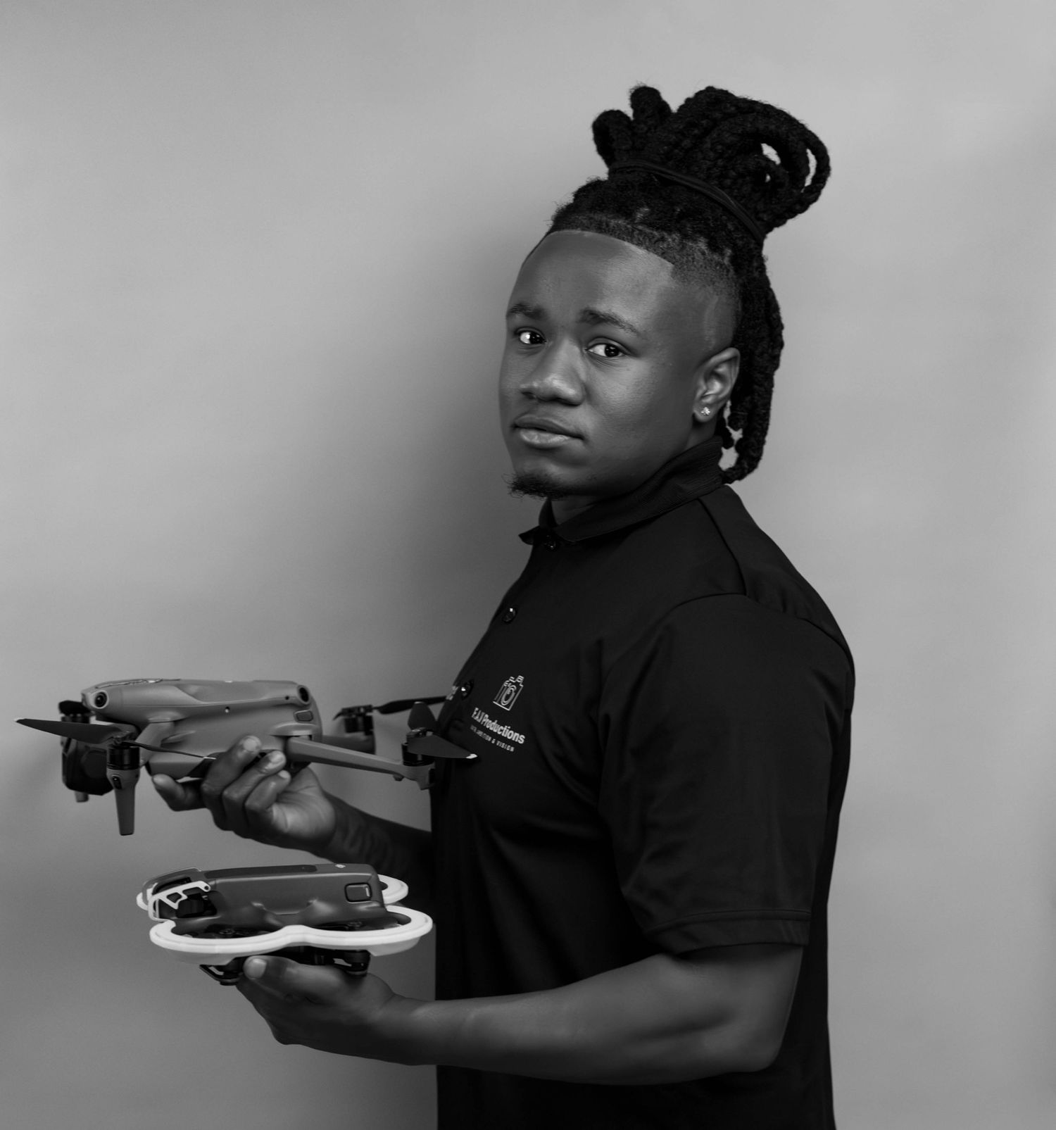 Black and white portrait of a young man with dreadlocks holding a drone, wearing a polo shirt.