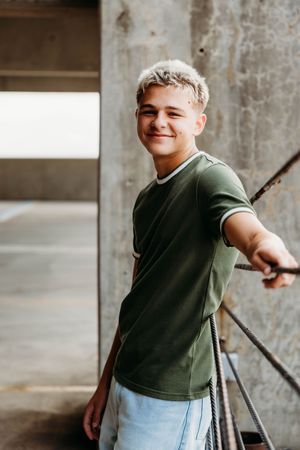 Smiling teenage boy with blonde hair wearing a green t-shirt, posing outdoors near concrete walls and metal railings.