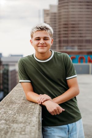 Young man with blonde hair in green ringer t-shirt leaning on rooftop ledge with city skyline behind him.