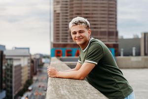 Young man with blonde hair leaning on rooftop ledge, smiling, with urban cityscape behind him.
