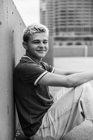 Black and white photo of a smiling teenage boy with blonde hair sitting against a wall in an urban setting.