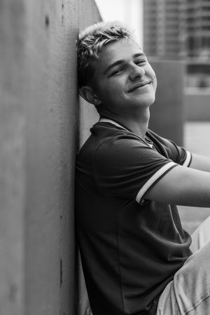 Black and white portrait of a smiling teenage boy leaning against a wall outdoors.