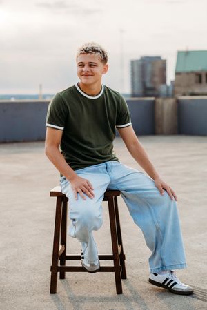 Young man sitting on wooden stool on rooftop, wearing green t-shirt and light jeans with prosthetic leg.