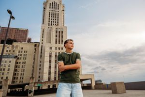 Young man standing on urban rooftop with arms crossed, tall building backdrop, wearing green t-shirt and jeans.