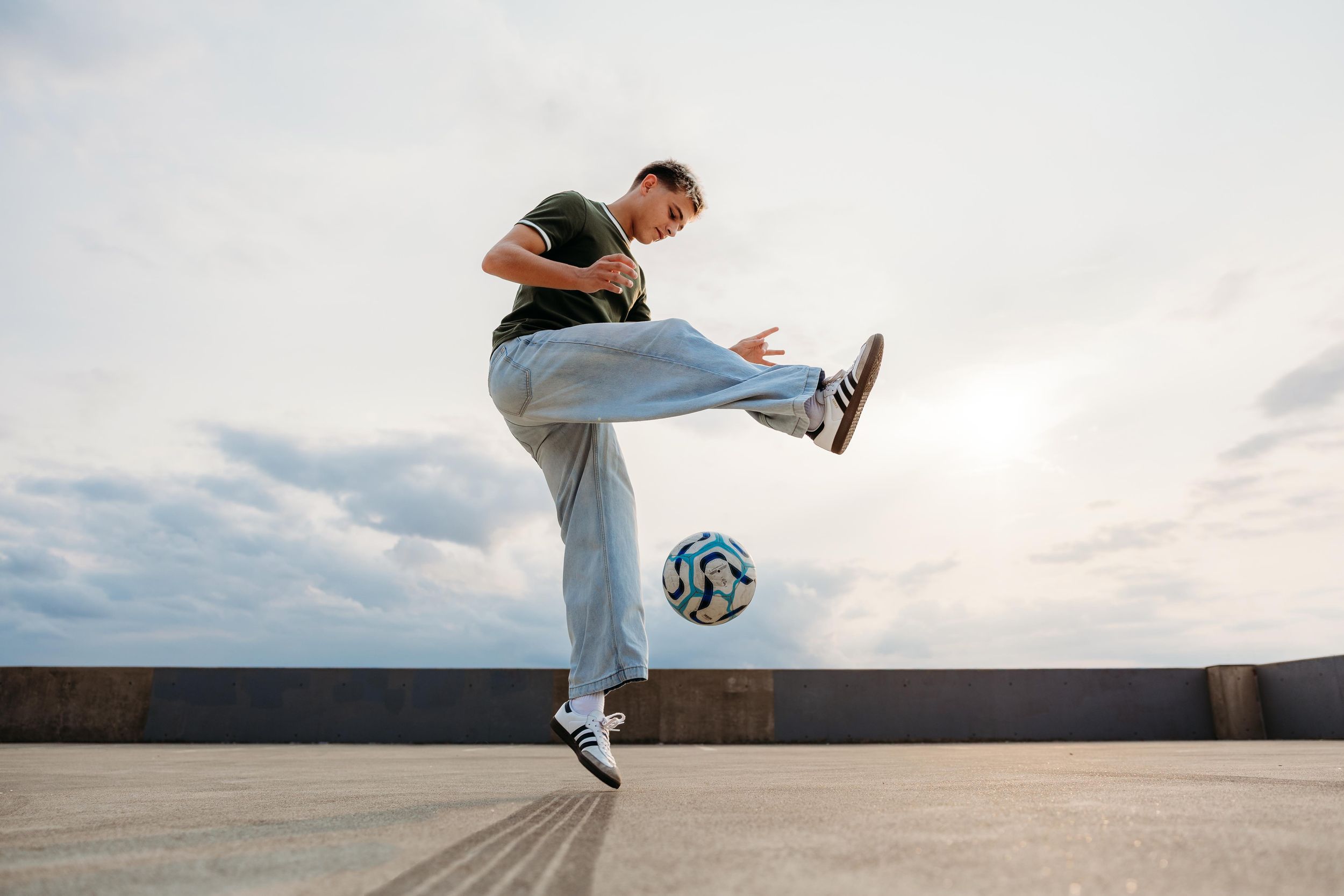 Young man performing freestyle soccer tricks, jumping mid-air while kicking a soccer ball outdoors under cloudy sky.