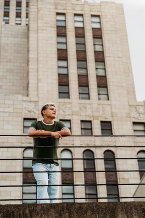 Young man in green t-shirt and jeans leaning on railing with urban building backdrop.