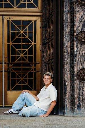 Young man in white shirt and jeans sitting relaxed against ornate Art Deco building entrance with gold doors.