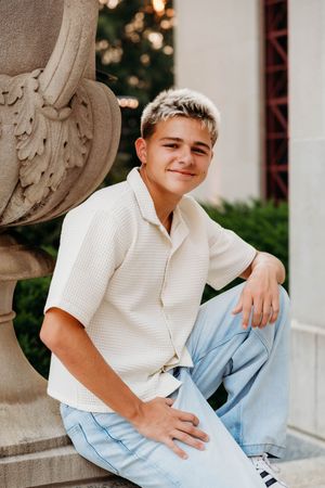 Young teen boy with blonde hair sits smiling outdoors near stone urn, wearing cream shirt and light blue jeans.