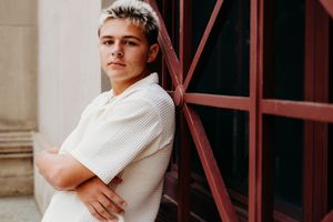 Young man with blonde hair in white polo shirt leaning against red window frame with arms crossed.