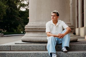 Young man in white polo and jeans sitting on stone steps near classical columns outdoors.