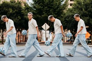 Four young men walking across a crosswalk with soccer balls in an urban setting, wearing white shirts and light blue jeans.