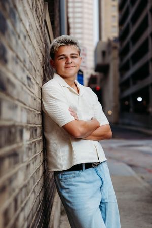 Young man with blonde hair leans against brick wall in urban alley, arms crossed, wearing white shirt and jeans.