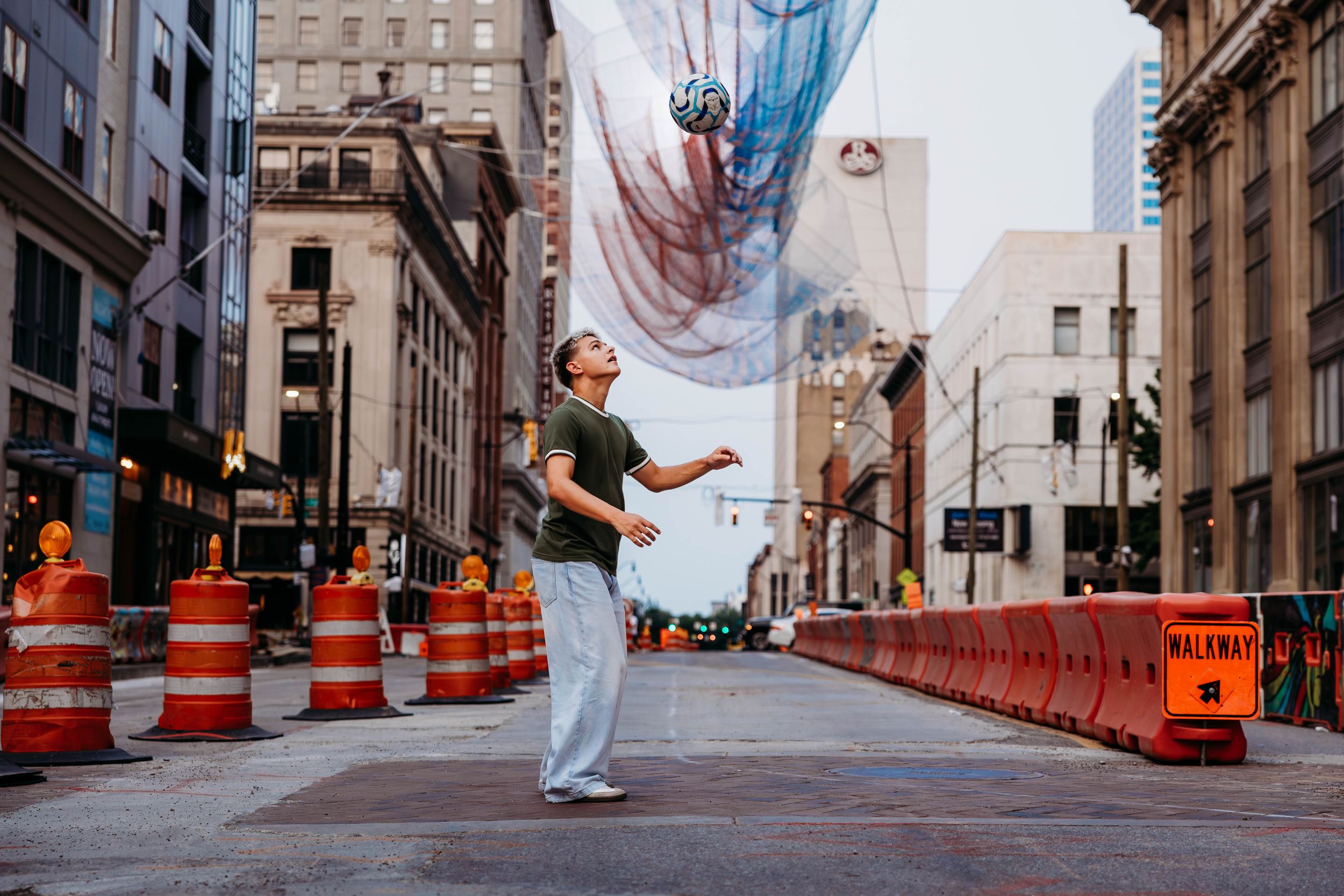 Man juggling a ball on an urban street with construction barriers and a colorful mural banner overhead.