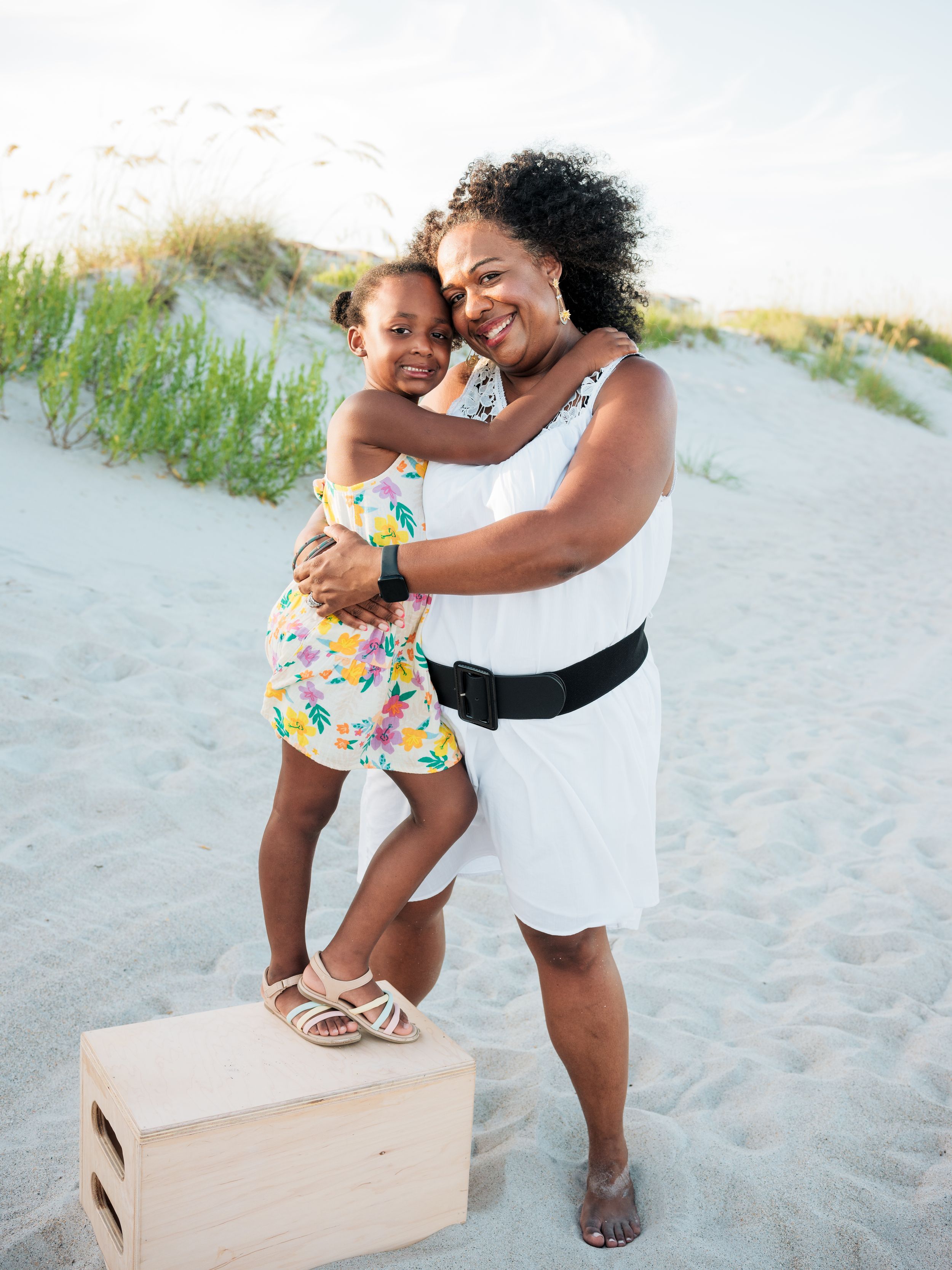A woman in a white dress hugs a young girl in a floral dress standing on a wooden box on a sandy beach.