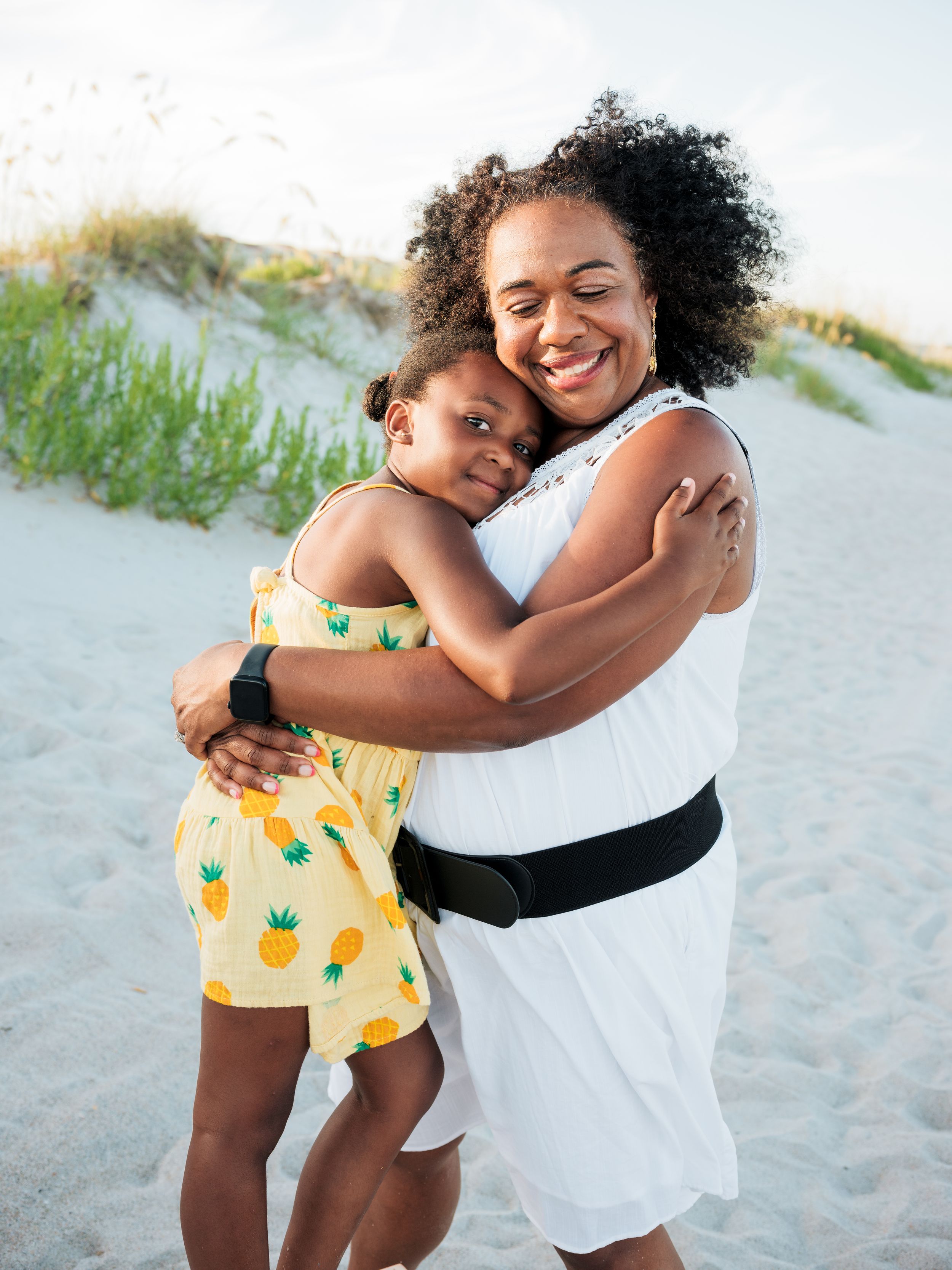 A smiling woman in a white dress hugs a young girl in a yellow pineapple dress on a sandy beach.